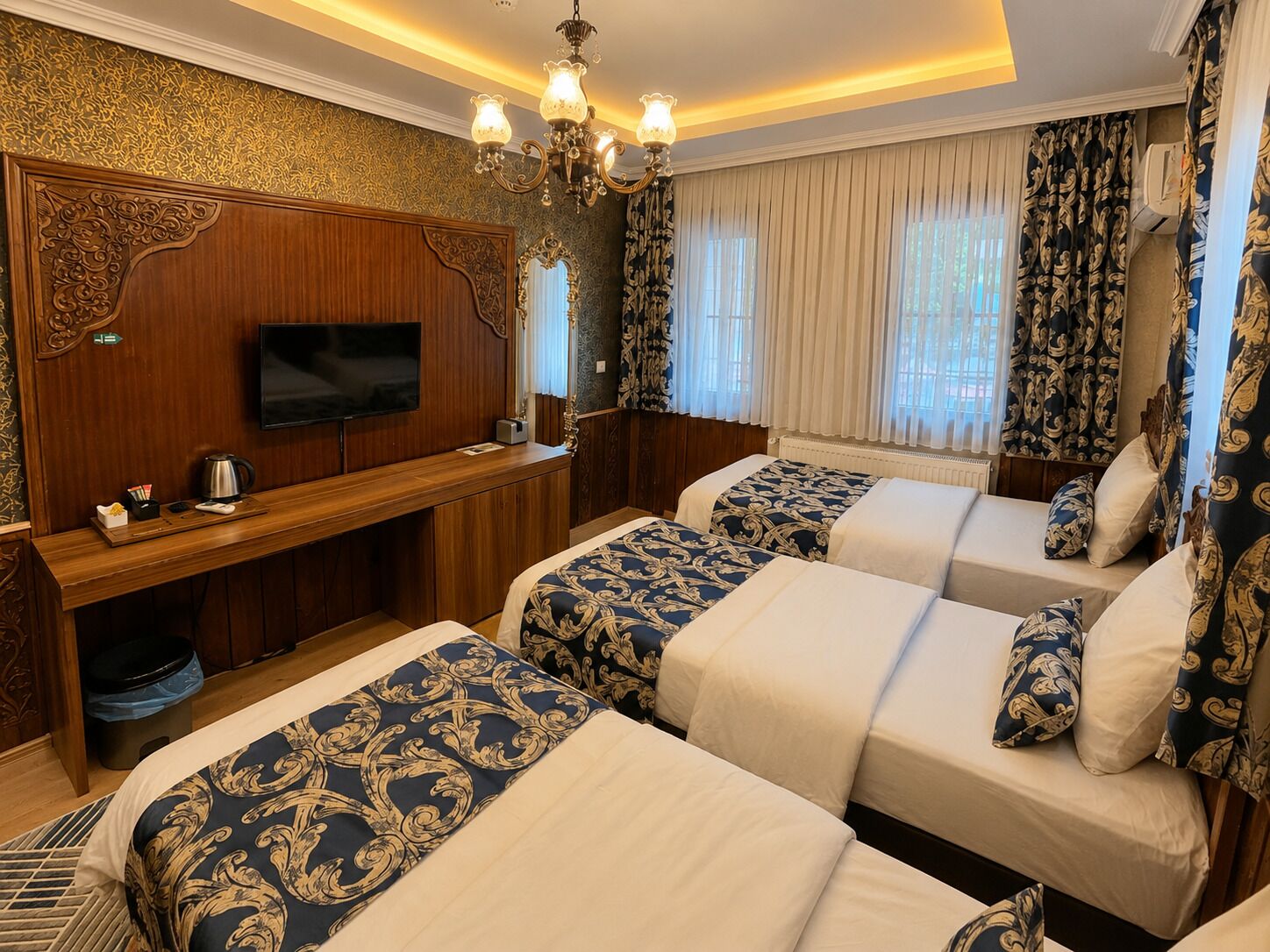 A hotel room with two beds, a TV, a desk with a kettle, and a chandelier.