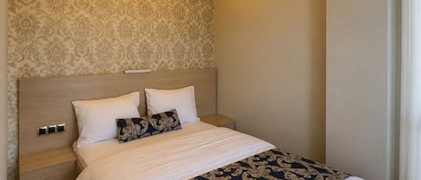 A modern bedroom with a patterned wallpaper, a wooden headboard, a bed with white and patterned bedding, and a bedside table with a lamp.