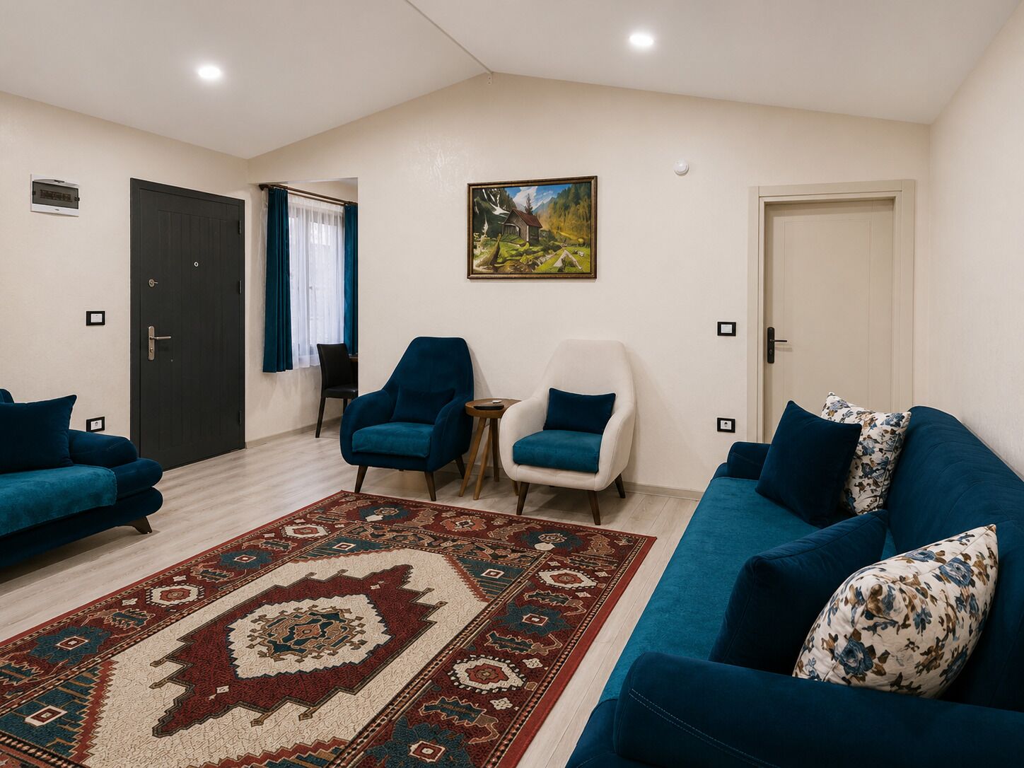 Ein modernes Wohnzimmer mit einem blauen Sofa, einem weißen Sessel und einem weiteren blauen Sessel mit blauem Kissen. Ein gemustertes Teppich liegt auf dem Boden, und ein Gemälde hängt an der Wand.