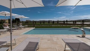 A pool area with lounge chairs and umbrellas, a clear sky, and a view of a landscaped area.