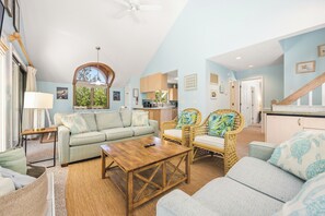 A spacious living room with a sofa, coffee table, and wicker chairs. A kitchen is visible in the background. - Sandy Paws: Dog-friendly, Corolla Light Amenities (Corolla)