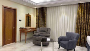 Quarto com porta de madeira, uma escrivaninha com espelho, uma poltrona cinza, uma mesa pequena e cortinas com padrão.