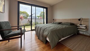 Un dormitorio con un ventanal grande, una cama con cabecero de madera, una silla y una mesita de noche con lámpara.