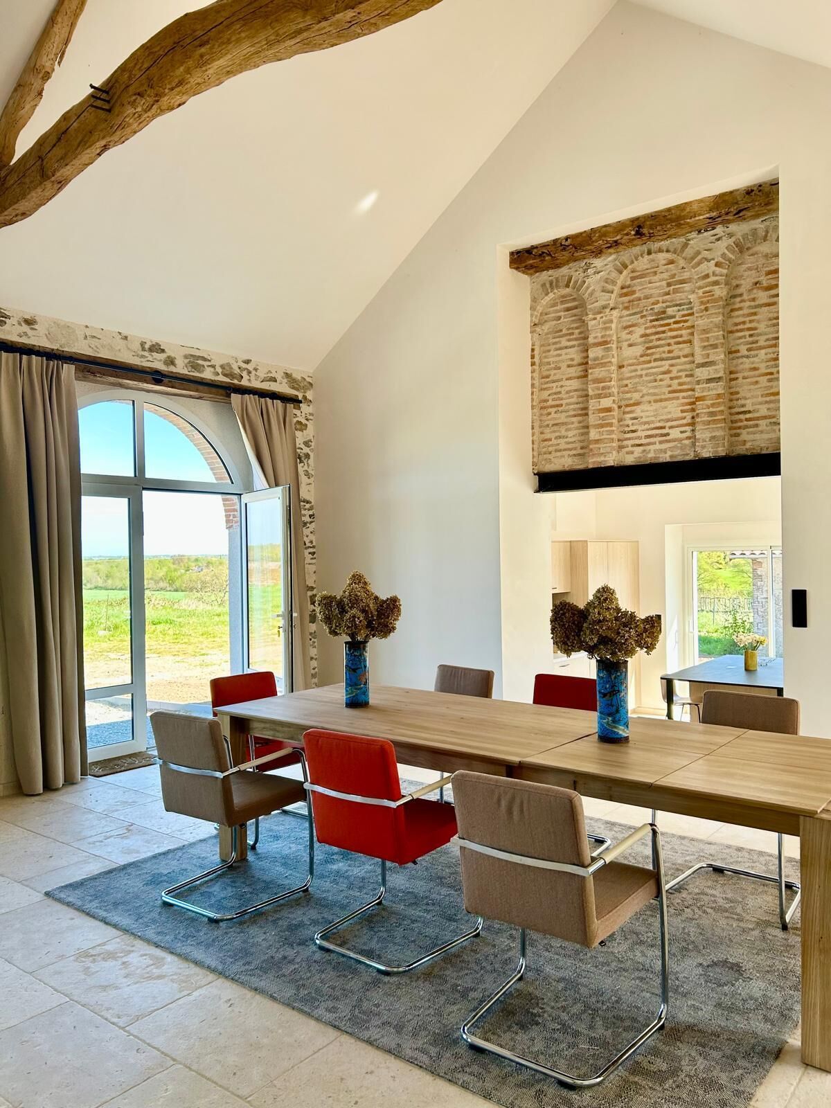 Une salle à manger moderne avec une longue table, des chaises rouges et beiges, et de grandes fenêtres.
