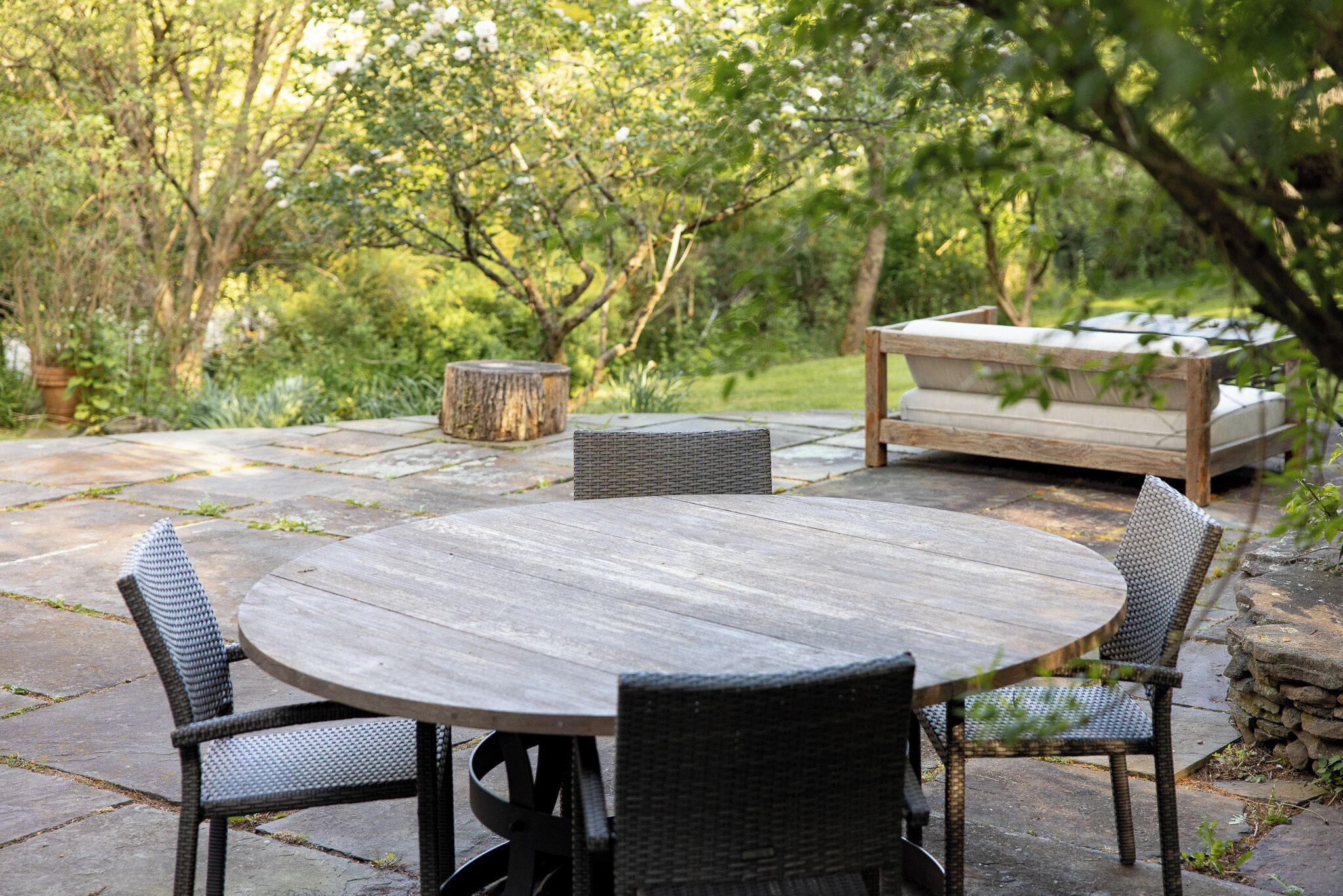 A round wooden table with wicker chairs, a stone bench, and a stone wall in a garden setting.