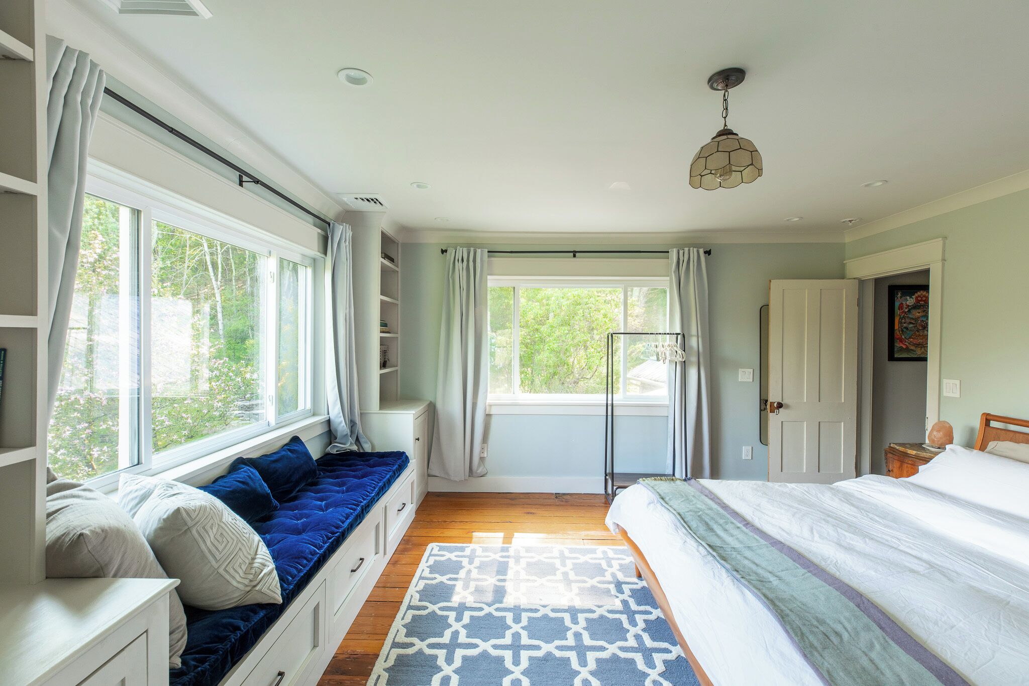 A bedroom with a large window, a bed, a built-in bench with cushions, and a patterned rug.