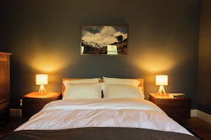 Une chambre d’hôtel avec un lit, deux tables de chevet, une petite table, une lampe et un tableau encadré au mur.