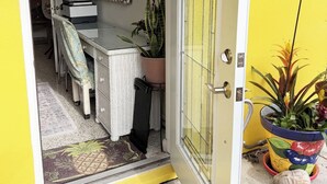 En gul yttervägg med en dörr som leder in till ett rum med ett matbord, stolar, en krukväxt och en spegel.