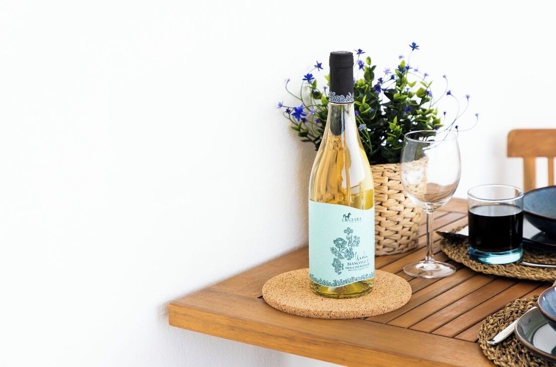 A bottle of white wine on a wooden table with a woven placemat, a glass, and a small vase with blue flowers.