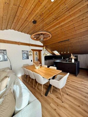 A modern interior with a wooden ceiling, a dining area with a wooden table and white chairs, a sofa, and a kitchen area. - Bergheimat Stüble Allgäu (Bad Hindelang)