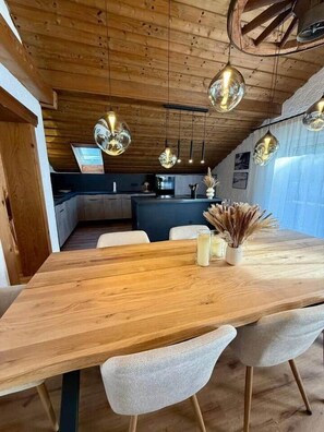 Ein Essbereich mit einem Holztisch, weißen Stühlen, hängenden Kugelleuchten und einer Küche im Hintergrund.