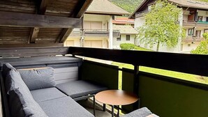 En balkon med sofa og lille bord, med udsigt over et bjergrigt område med bygninger.