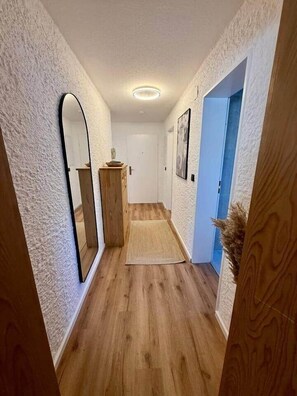 Ein Korridor mit Holzdielen, weißen, strukturierten Wänden, einem Spiegel, einem Holzkasten und einer blauen Tür.