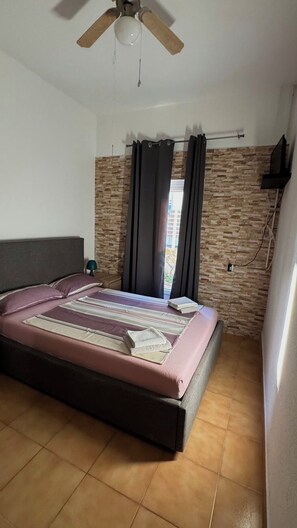 A bedroom with a bed, a ceiling fan, a window with curtains, a brick wall, and a television mounted on the wall. - Directly on the beach (Vir)