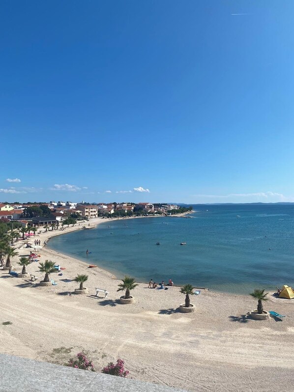 A coastal beach scene with clear skies, a sandy beach, palm trees, and buildings in the background. - Directly on the beach (Vir)