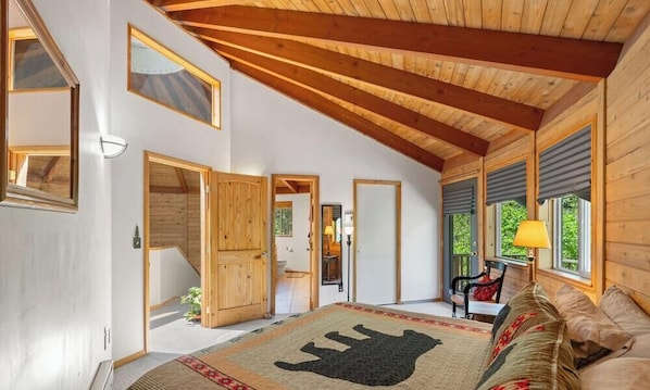 Ein geräumiges Zimmer mit hoher Decke, Holzbalken, einem großen Bett und einem Teppich mit Muster, mit Blick auf viel Grün.