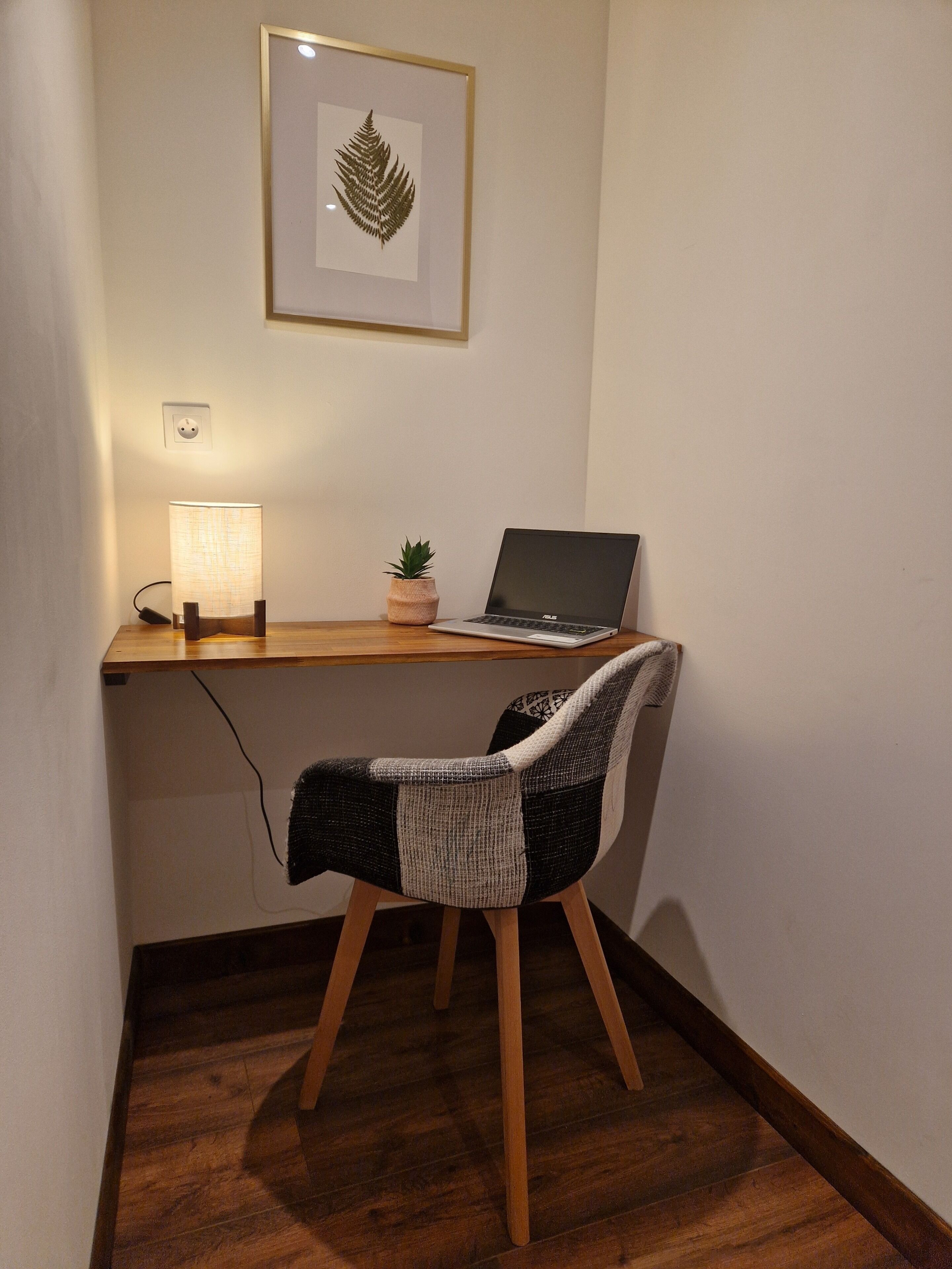 Een werkplek met een laptop, een lamp, een kamerplant en een lijst met een blad erop.