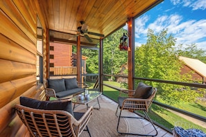 A wooden cabin with a balcony, wicker furniture, and a view of the surrounding forest. - Cozy 2-bedroom cabin in Gatlinburg with hot tub, game loft, pool (Gatlinburg)