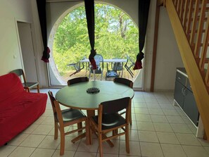 Ein Essbereich mit rundem Tisch und Stühlen, einem roten Sofa und Blick durch ein großes rundes Fenster auf eine Terrasse.