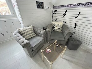 Ein Wohnzimmer mit grauem Sofa, passendem Sessel, einem kleinen Tisch und einem grauen Hocker. An den Wänden befinden sich Dekorationen, darunter ein Poster mit 'Rock & Roll' und eine Wand mit Noten.