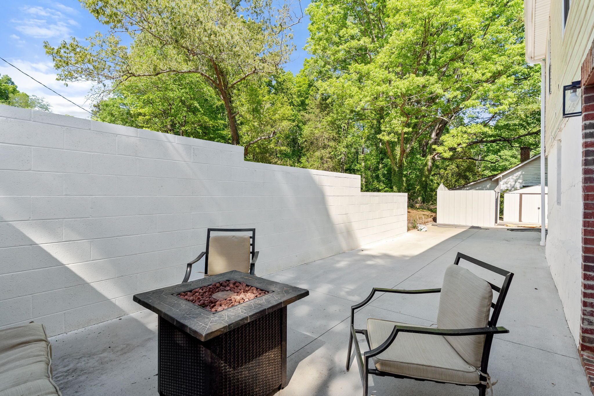 An outdoor patio with a fire pit table, two chairs, and a white brick wall.