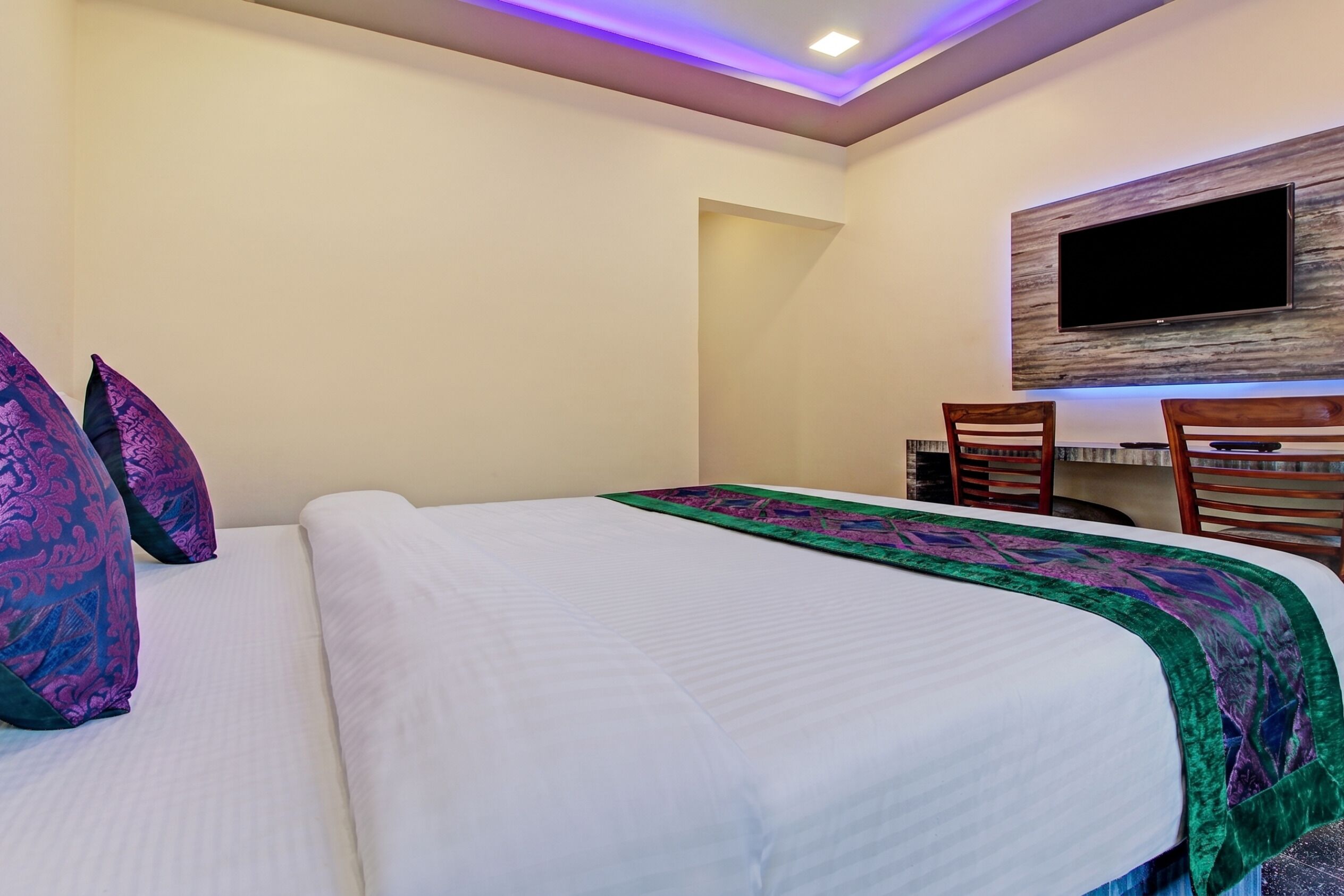 A hotel room with a bed, a desk with chairs, a flat-screen TV mounted on the wall, and a ceiling light.