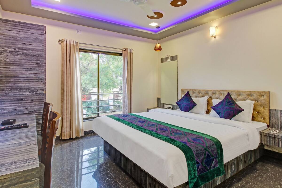 A hotel room with a large bed, a window with curtains, a bedside table, a nightstand, a lamp, and a ceiling light.