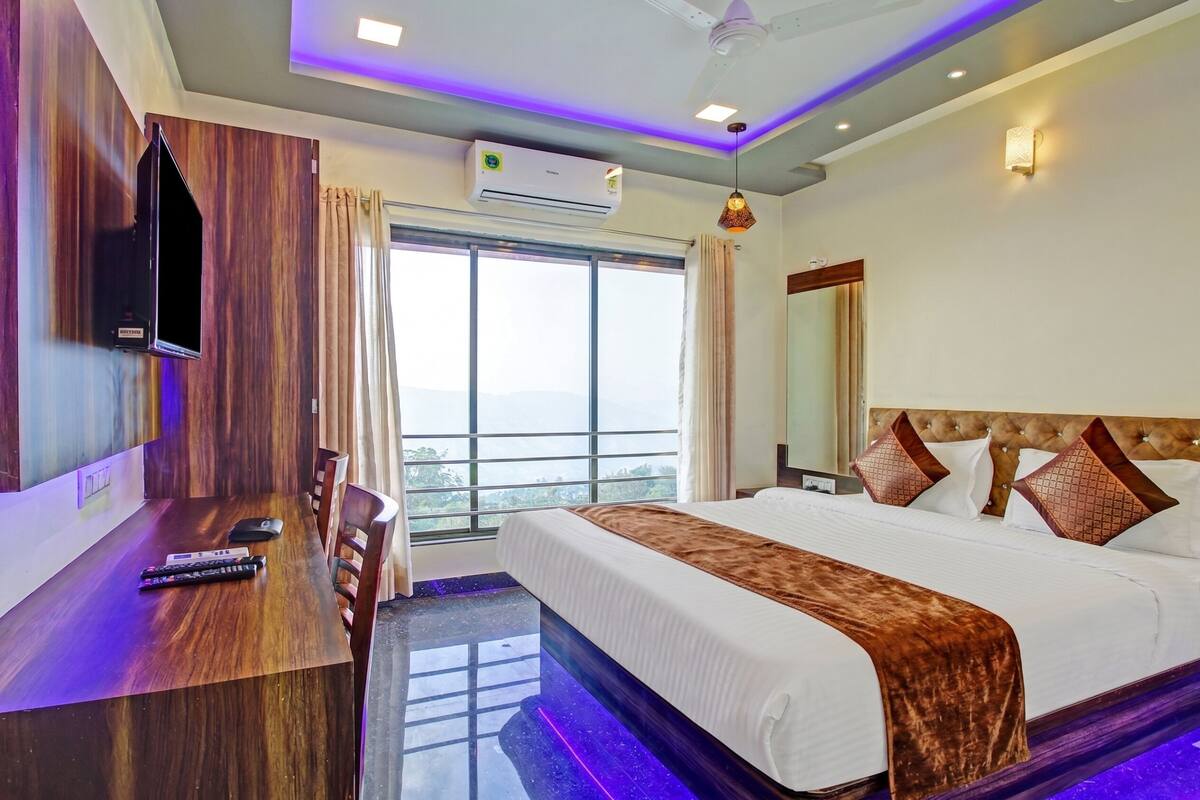 A hotel room with a large bed, a desk with a remote control, an air conditioner, and a view of the outdoors.