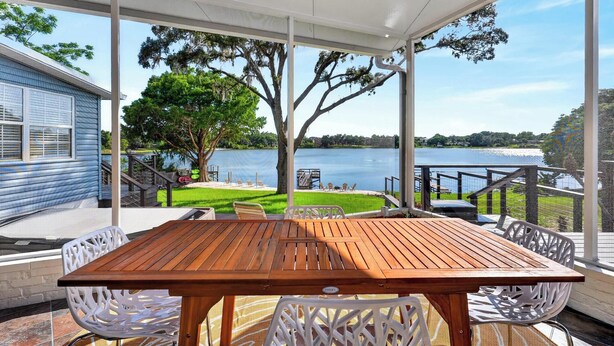 A covered patio with a wooden table and white chairs, overlooking a lake with a grassy area and a dock.