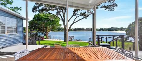A covered patio with a wooden table and white chairs, overlooking a lake with a grassy area and a dock.