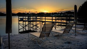 A beach with lounge chairs and a wooden deck overlooking a calm lake at sunset.