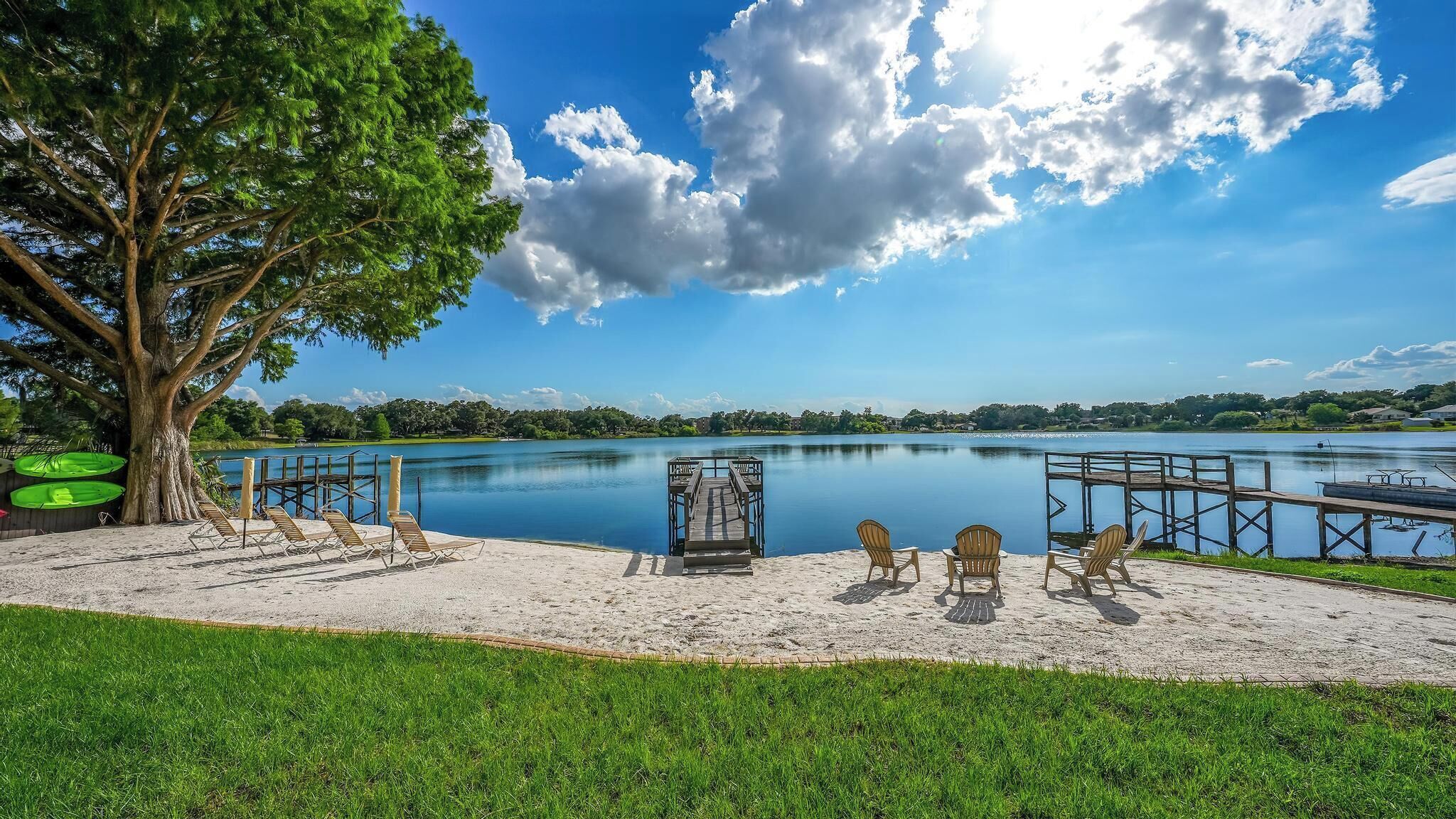 A lakeside view with a dock, chairs, and a large tree.