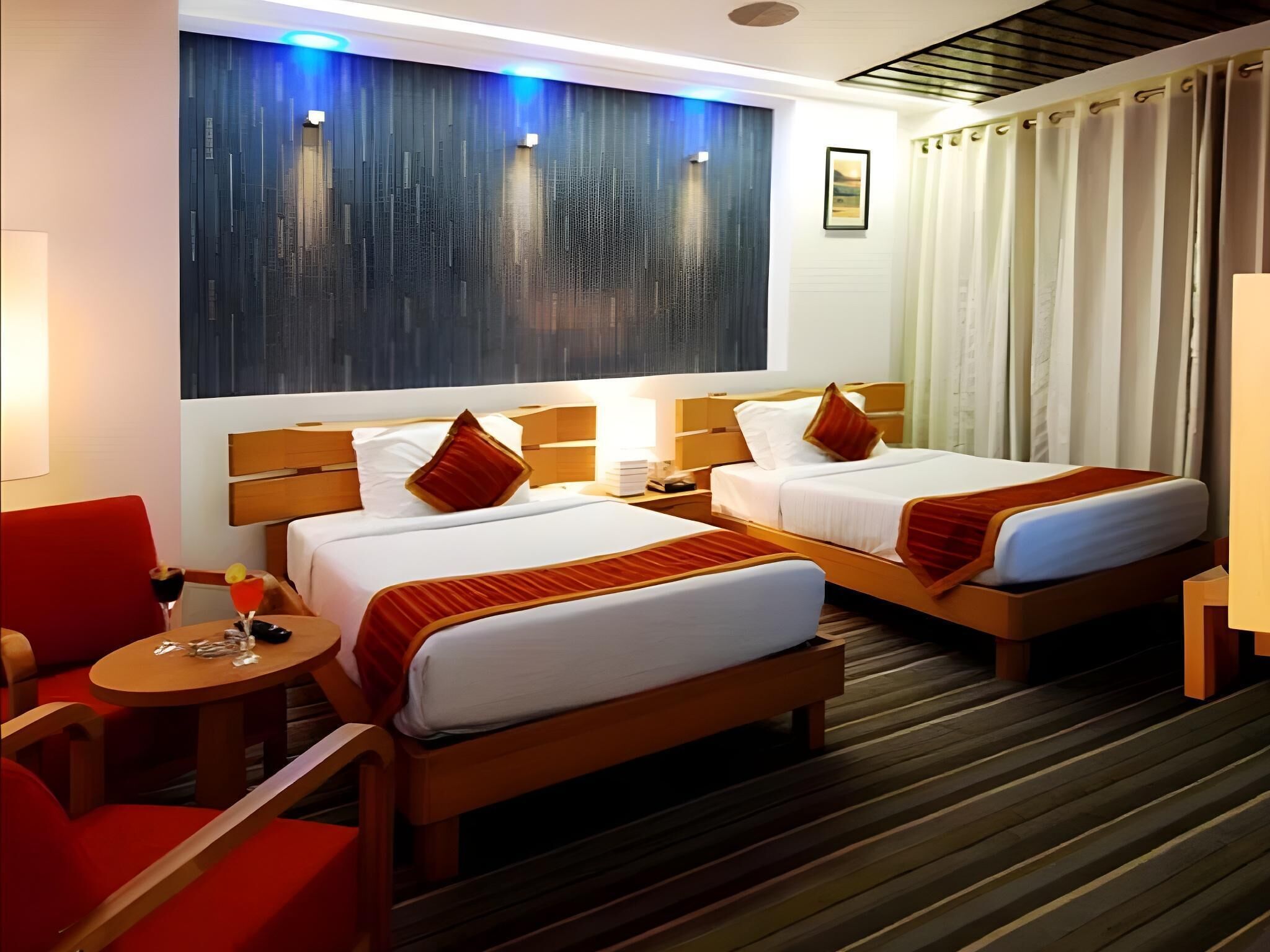 Quarto de hotel com duas camas, uma cabeceira de madeira, uma mesa redonda pequena, uma cadeira vermelha e uma obra de arte pendurada na parede.