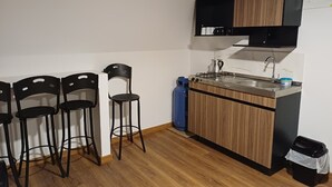 Cocina compacta con estufa a gas, lavamanos y armarios de madera. Hay tres taburetes negros y una silla. Se observa un cilindro de gas azul.