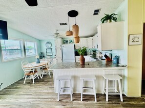A kitchen with a central island, white cabinetry, a ceiling fan, and a dining area with white chairs. - Ocean Breeze Retreat w/ Dock - Keys Escape (Cudjoe Key)
