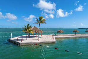 A beachside pier with thatched-roof structures, palm trees, and a clear blue sky. - Ocean Breeze Retreat w/ Dock - Keys Escape (Cudjoe Key)