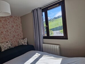 A bedroom with a bed, floral wallpaper, a window with a view of a green landscape and mountains, and a radiator. - Cosy cocoon with mountain views (5 pers) at the foot of the slopes and paths (Le Grand-Bornand)