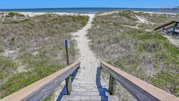 Ein hölzerner Steg führt zu einem Strand mit Dünen und vereinzelter Vegetation.