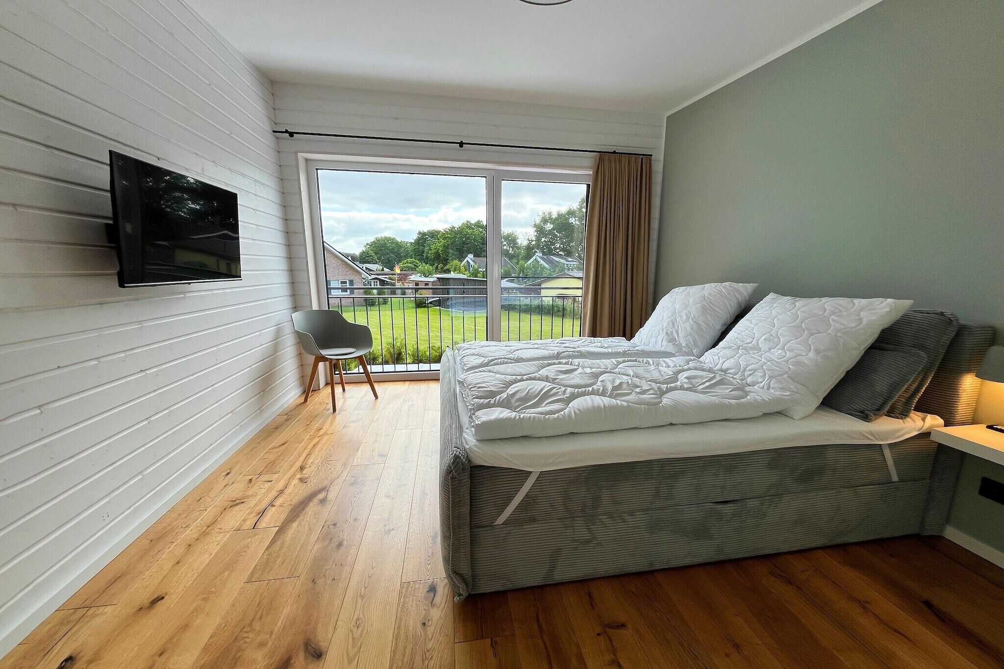 Une chambre à coucher comprenant un lit, une chaise, un téléviseur fixé au mur et une grande fenêtre donnant sur l’extérieur.