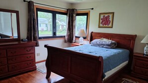 A bedroom with a wooden bed, a dresser, a nightstand, a window with curtains, and a wall-mounted artwork. - Charming 1-bedroom available in the house  Burlington perfect for a cozy getaway (Burlington)