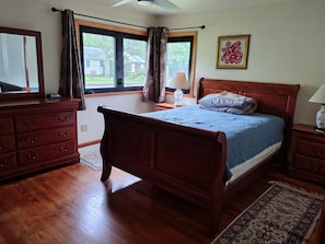 Une chambre à coucher avec un lit en bois, une commode, une table de chevet, une fenêtre avec des rideaux et une lampe fixée au mur.