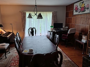 Un espace repas comprenant une longue table, des chaises, un piano, un bureau avec un ordinateur et un téléviseur.