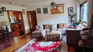 A living room with a sofa, ottoman, and a red and white patterned rug. There is a ceiling fan, a dining area with a table and chairs, and a kitchen visible through an open door. - Charming 1-bedroom available in the house  Burlington perfect for a cozy getaway (Burlington)