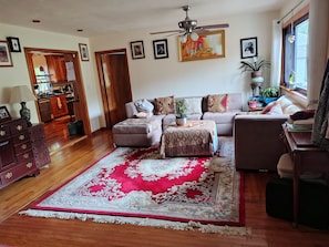 Un salon avec un canapé, un pouf et un tapis rouge et blanc. On y trouve un ventilateur de plafond, un coin repas avec une table et des chaises, et une cuisine visible à travers une porte ouverte.