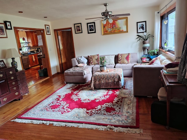 A living room with a sofa, ottoman, and a red and white rug. There is a ceiling fan, a dining area with a table and chairs, and a kitchen visible through an open door.