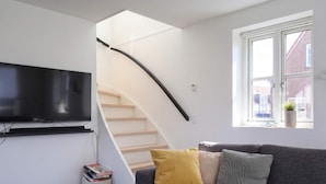 A modern living room with a grey sofa, a flat-screen TV mounted on the wall, a staircase, and a window with a view of a building outside. - 4 Star Holiday Home in Lokken (Lokken)