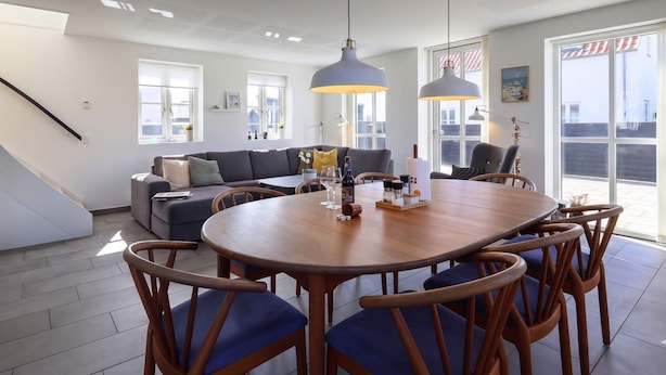 A modern living room with a wooden dining table, blue chairs, a grey sofa, and large windows. - 4 Star Holiday Home in Lokken (Lokken)