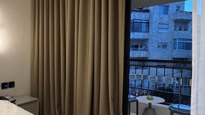 A hotel room with a bed, curtains, a balcony with a table and chairs, and a view of buildings. - 309 Tara-jre 2bedroom at Jabal Al Lweibdeh (Amman)