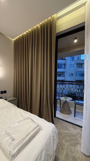 A hotel room with a bed, curtains, a balcony with a table and chairs, and a view of buildings. - 309 Tara-jre 2bedroom at Jabal Al Lweibdeh (Amman)