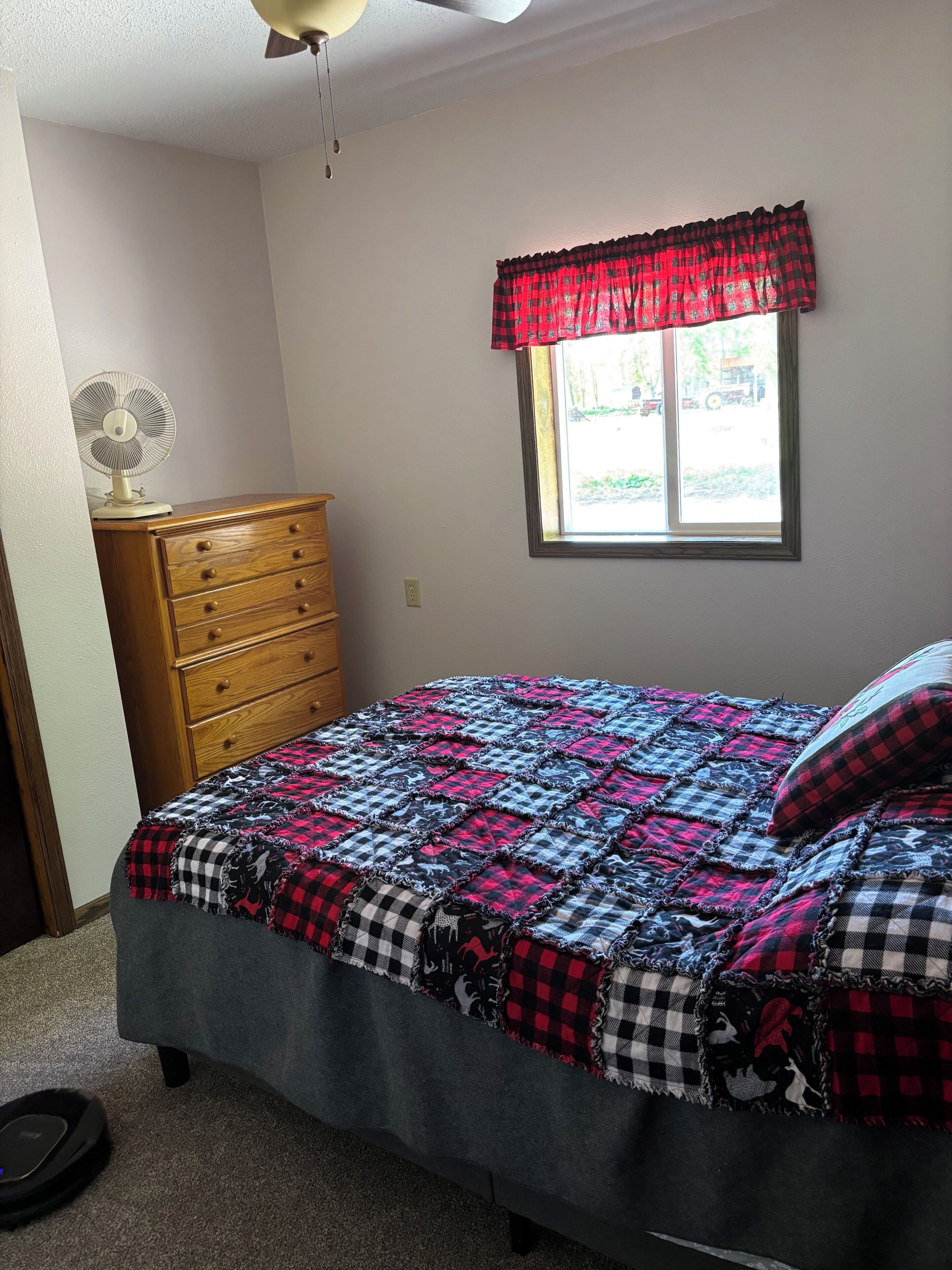 A bedroom with a bed, a dresser, a window with a red valance, and a ceiling fan.